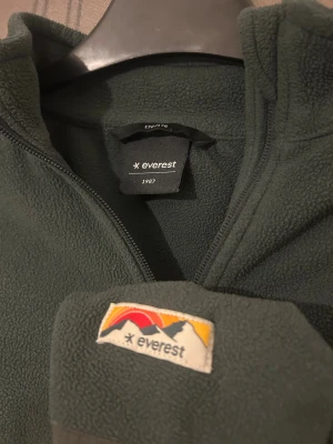 Mörkgrön fleece från Everest - Mysig mörkgrön fleece från Everest med hel dragkedja. Fleecen har ett broderat Everest-märke på ärmen och är perfekt för kalla dagar. Enkel och stilren design med lång ärm och mjukt material som håller dig varm.