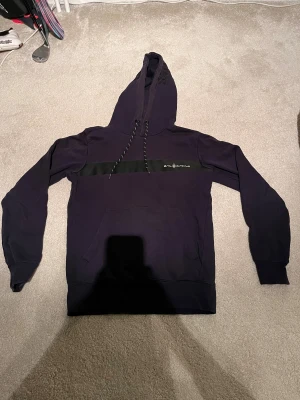 Mörkblå Sail Racing hoodie i stl 170  - Snygg mörkblå hoodie från Sail Racing i storlek 170. Tröjan har en bred svart rand över bröstet med logga, snörning i huvan och en stor magficka. Materialet är mjuk bomull och polyester, perfekt för chill dagar. Diskret Sail Racing-tryck på huvan för extra stil.