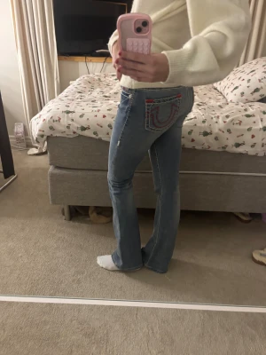 True Religion blå bootcut jeans - Säljer ett par blå jeans från True Religion med ikonisk röd och vit kontrastsöm och broderad logga på bakfickan. Modellen har bootcut-ben. Materialet är slitstark denim i bomull. Använd ett par fåtal gånger och därmed är priset högt, köpte jeansen för 2200.