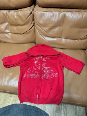 Röd hoodie från costa nostra med ”diamanter” - Cool röd hoodie med korta ärmar, huva och hel dragkedja framtill. Framsidan har ett stort tryck med glittriga detaljer och text. Mjuk insida i fleece som gör den extra skön. Perfekt för dig som vill sticka ut med en unik streetstyle.