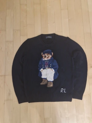 Mörkblå stickad tröja Polo Ralph Lauren - Mörkblå stickad tröja från Polo Ralph Lauren med ikonisk björn på framsidan. Tröjan har rund halsringning, ribbade muddar och RL-broderi nedtill. Perfekt för dig som gillar klassisk preppy stil och vill sticka ut med en cool detalj.