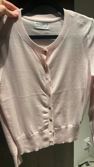 Ljusrosa kofta från Holly & Whyte - Supersöt ljusrosa kofta från Holly & Whyte med rund halsringning och guldiga knappar framtill. Långärmad och i mjukt, tunt material som känns skönt mot huden. Perfekt att slänga över en topp när det är lite kyligt ute.