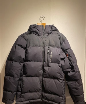 Sail Racing jacka - Svart pufferjacka från Sail Racing i storlek S. Jackan har huva, flera fickor med dragkedja och en clean design med Sail Racing-logga på bröstet. Perfekt för kalla dagar med sin tjocka vaddering. Är i mycket bra skick och använd fåtal gånger.