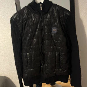 Svart quiltad jacka Emporio Armani - Svart jacka från Emporio Armani med quiltad framsida i blankt material och stickade ärmar och rygg. Jackan har dragkedja framtill, ribbad krage och muddar samt en snygg logotyp på bröstet. Perfekt för dig som vill ha en stilren och exklusiv look.