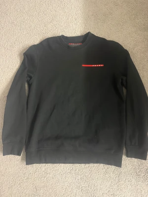 Svart sweatshirt från Prada, storlek L - Svart sweatshirt från Prada i storlek L med diskret röd logga på bröstet. Tröjan har rund hals och långa ärmar. Tillverkad i 90% bomull och 10% elastan för en skön och mjuk känsla. Perfekt för dig som gillar stilrena designer och exklusiva märken.