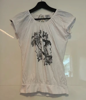 Vit blus med svart konsttryck b.young - Vit blus från b.young med korta ärmar och rund halsringning med knyt i nacken. Framsidan har ett svart konstnärligt tryck med ansikte och blommor. Blusen är lätt rynkad upptill och har bredare kant nertill. Perfekt för dig som gillar unika detaljer. Även insydda i kanten på magen, så sitter som en xs