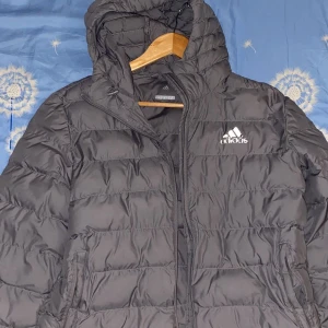 Svart pufferjacka från Adidas - Svart pufferjacka från Adidas med huva och dragkedja framtill. Jackan har quiltad design och Adidas-logga på bröstet. Perfekt för kyliga dagar och har en sportig vibe.