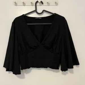 Svart croppad blus från Shein - Svart croppad blus från Shein med djup v-ringning och vida trekvartsärmar. Toppen har smockade partier i midjan som ger en snygg passform och flowig känsla. Perfekt för dig som gillar trendiga och stilrena plagg.