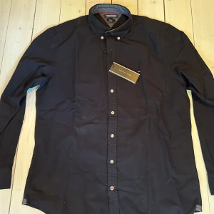Aldrig använd Mörkblå skjorta Tommy Hilfiger slim fit - Mörkblå långärmad skjorta från Tommy Hilfiger i slim fit-modell med stretch. Klassisk krage, vita knappar och diskret logga vid bröstet. Skjortan är i bomull och har en stilren look som passar perfekt till jeans eller chinos. Har aldrig blivit använd - helt ny.