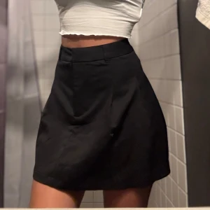 Svart kortkjol med klassisk passform - Enkel och stilren svart kortkjol med hög midja och lätt utsvängd form. Kjolen har slät framsida och diskreta sömmar, perfekt för att matcha med crop tops eller stickade tröjor. Passar dig som gillar minimalistisk och clean stil.