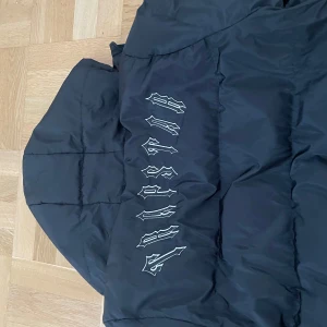 Svart pufferjacka från Urban Classics - Svart pufferjacka med huva från Urban Classics. Jackan har en stor logga i vitt på ryggen och en rund patch på bröstet. Dragkedja framtill och två fickor med tryckknapp. Perfekt för kalla dagar och riktigt street i stilen.