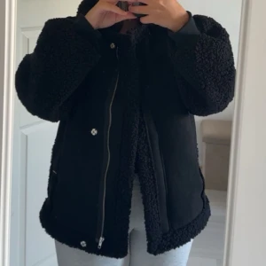 Svart teddyjacka med dragkedja - Mysig svart teddyjacka med fluffiga ärmar och detaljer längs kanterna. Jackan har både dragkedja och knappar framtill, samt en loose passform som ger en chill vibe. Perfekt för kalla dagar när du vill vara både varm och snygg.