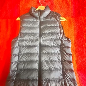 Svart Marinblå dunväst från Uniqlo XS - Säljer en svart, ultralätt dunväst från Uniqlo i storlek XS. Västen har quiltad design, hög krage och dragkedja framtill. Tillverkad i syntetmaterial med dunfyllning. Perfekt för lager-på-lager och enkel att packa ner i medföljande påse.