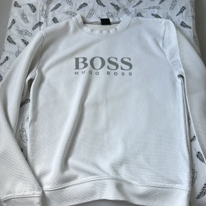 Vit Hugo Boss sweatshirt - Vit långärmad sweatshirt från Hugo Boss med stor logga framtill. Tröjan har en strukturerad yta och rund halsringning. Perfekt för dig som gillar stilrena och klassiska plagg med en modern touch.