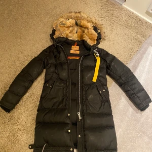 Svart Parajumpers dunjacka med päls XS i bra skick men andvänd några år 2500 kr - Säljer en svart lång dunjacka från Parajumpers i storlek XS. Jackan har en fluffig huva med äkta päls, gul detalj vid dragkedjan och flera fickor framtill. Perfekt för kalla dagar och riktigt snygg streetstil.