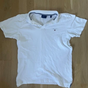 Vit pikétröja från GANT, storlek S - Vit pikétröja från GANT med klassisk krage och broderad logga på bröstet. Tröjan har korta ärmar och blå randig detalj på insidan av kragen. Tillverkad i bomull för en skön och luftig känsla.