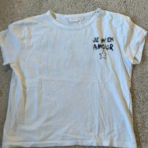 Vit t-shirt med tryck från Gina Tricot - Snygg vit t-shirt från Gina Tricot med svart texttryck 'JE M'EN AMOUR' och en stjärna både fram och bak. Modellen är klassisk med rund hals och korta ärmar, tillverkad i mjuk bomull. Perfekt för dig som gillar coola statement-plagg.