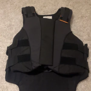 Svart säkerhetsväst  - Säljer en svart säkerhetsväst från Airowear, modell Teen Outlyne. Västen har justerbara kardborreband på sidorna och axlarna samt orange detaljer. Perfekt för ridning och ger bra skydd. Materialet är syntet och västen är designad för att sitta bekvämt och säkert.