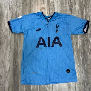 Blå Tottenham Hotspur fotbollströja Nike - Snygg blå Tottenham Hotspur fotbollströja från Nike med mörkblå detaljer och klubbmärke på bröstet. Tröjan har korta ärmar, diskret mönster i tyget och AIA som sponsortryck på framsidan. Tillverkad i lätt och ventilerande material.