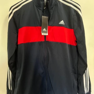 Adidas Kofta med röd detalj XL - Snygg kofta från Adidas i mörkblått med en bred röd panel över bröstet och vita ränder längs axlarna. Jackan har dragkedja framtill, ribbad krage och klassisk Adidas-logga på bröstet. Tillverkad i polyester och Primegreen-material. Oanvänd