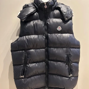 Svart dunväst från Moncler, storlek L - Snygg svart dunväst från Moncler med NFC Märke och med quiltad design och två dragkedjefickor framtill. Västen har hög krage och klassisk Moncler-logga på bröstet. Insidan har en cool serietidningsdetalj. Perfekt för lager-på-lager-stil under kyliga dagar.