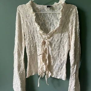 Vit spetsblus med snörning och volang - Romantisk vit blus från Gina tricot i spets med långa ärmar, volangkant längs v-ringningen och snörning framtill. Blusen är lätt transparent och fint spetsmönster