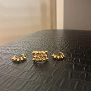 Edblad Guldiga örhängen och ring med nitar - Edblad set med två guldiga örhängen och en matchande ring, alla dekorerade med små nitar för en edgy vibe. Snyggt och unikt smycke som sticker ut och passar perfekt för dig som gillar statement-accessoarer.