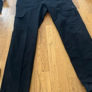 Svarta cargopants i bomull - Svarta cargopants med raka ben och stora fickor på sidorna. Byxorna är tillverkade i 100% bomull och har en klassisk look med knappstängning på fickorna. Perfekta för en avslappnad och stilren outfit.