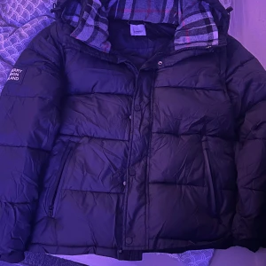 Svart pufferjacka från Burberry - Svart pufferjacka från Burberry med stor logga på ärmen och klassiskt rutigt foder i huvan. Jackan har dragkedja, knappar och två fickor framtill. Perfekt för kalla dagar och riktigt snygg streetstil.