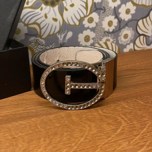 Svart skärp med strass och stor logga Guess - Snyggt svart bälte från Guess i glansigt skinn med bred design. Spännet är stort, ovalt och pryds av strass-stenar samt Guess-logga i silver. Insidan är ljus och har Guess-stämpel. Perfekt för att lyfta din outfit med extra bling.