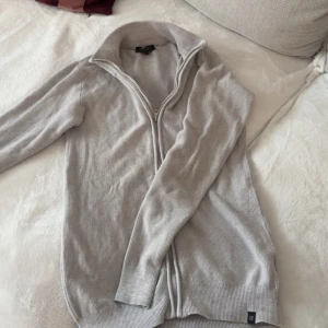 Ljusgrå stickad kofta med dragkedja - Ljusgrå stickad kofta med hel dragkedja framtill och hög krage. Perfekt för lager-på-lager och enkel att matcha med olika outfits. Långärmad och mjuk i materialet för extra komfort.