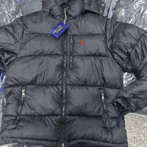 Svart pufferjacka från Polo Ralph Lauren - Svart pufferjacka från Polo Ralph Lauren med röd broderad logga på bröstet. Jackan har huva, flera dragkedjefickor och quiltad design. Perfekt för kalla dagar och har en modern, sportig look.