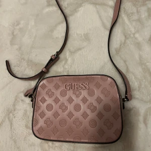 Rosa axelväska från Guess i skinn - Snygg rosa axelväska från Guess i skinnimitation med präglat logomönster över hela väskan. Justerbar axelrem och dragkedja upptill. Insidan är ljus med Guess-logga och har ett innerfack med dragkedja. Perfekt storlek för det viktigaste.
