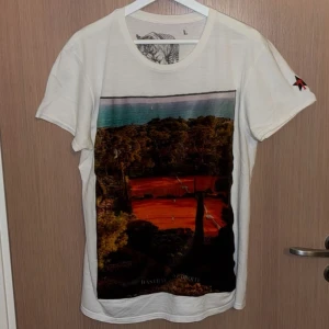 Vit Bastille t-shirt - Vit t-shirt från Bastille i bomull med stort färgglatt tryck av tennisbana och natur på framsidan. På ena ärmen finns en svart stjärna med röd palm. Klassisk passform och rund hals. Perfekt tisha för sommardagar. Storlek L men sitter som M
