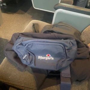 Blå Djurgår'n midjeväska - Säljer en blå midjeväska från järnkaminerna med broderad logga framtill. Väskan har flera dragkedjefack och justerbart band, perfekt för att bära det viktigaste. Enkel och sportig design som funkar till många tillfällen.