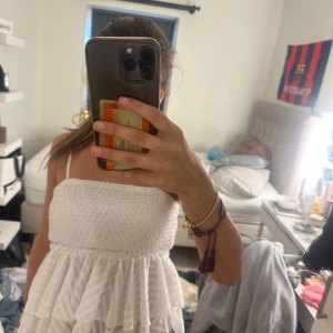 Vit somrig kortklänning med volanger - Supersöt vit kortklänning med smockad överdel och tunna axelband. Kjolen har flera lager volanger som ger en luftig och lekfull känsla. Perfekt för varma dagar och har en riktigt fräsch vibe. Materialet känns lätt och bekvämt mot huden.