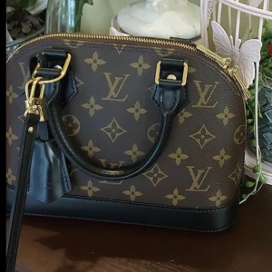 Louis Vuitton Monogram handväska - Snygg handväska från Louis Vuitton i klassiskt monogrammönster. Väskan är brun med svarta detaljer och guldfärgade metalldelar. Tillverkad i skinn med dragkedja upptill och två handtag. Perfekt för dig som vill ha en ikonisk accessoar.