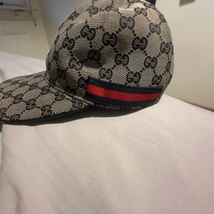 Gucci keps med GG-mönster och röd-blå rand - Snygg keps från Gucci med klassiskt GG-monogram i beige och svart. Kepsen har en röd och marinblå rand på sidan och justerbar rem baktill i svart skinn. Tillverkad i bomullscanvas med detaljer i läder. Perfekt för dig som vill sticka ut med en lyxig accessoar.