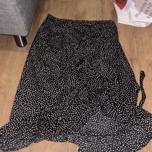 Svart prickig omlottkjol Vero Moda - Svart omlottkjol från Vero Moda med vita prickar och volangkant. Kjolen har knytband i sidan 