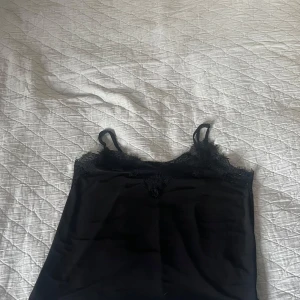 Svart linnetopp med spetsdetaljer XS - Säljer en svart linnetopp i storlek XS med tunna axelband och fina spetsdetaljer längs urringningen. Toppen har en slät, mjuk yta och är perfekt för att skapa en stilren look. Tillverkad i ett lätt material som känns skönt mot huden.