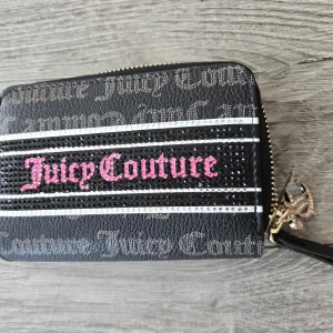 Juicy Couture svart plånbok med strass - Svart plånbok från Juicy Couture med glittriga strassdetaljer och rosa logga framtill. Plånboken har dragkedja runtom, flera kortfack och fack för mynt. Snyggt mönstrad insida med Juicy Couture-tryck. Perfekt accessoar för dig som gillar bling och streetstyle.