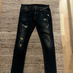 Mörkblå skinny jeans från Nudie Jeans - Snygga mörkblå jeans från Nudie Jeans med slitna detaljer och orange sömmar på bakfickorna. Modellen är skinny fit och har klassisk femficksdesign. Jeansen är tillverkade i bomull och har en cool, lite sliten look. 31/30