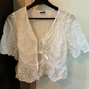 Vit blus med broderade volanger - Superfin vit blus med korta ärmar, broderade volangdetaljer och snörning framtill. Blusen har v-ringning och är croppad, perfekt för sommarens alla tillfällen. Tillverkad i bomull för en luftig känsla.