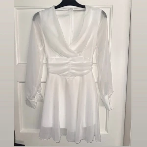 Vit omlottklänning med v-ringning - Säljer en vit kortklänning med omlottliv och v-ringad hals. Klänningen har långa, transparenta ärmar med knappar vid mudden och ett markerat midjeparti med rynkade detaljer. Materialet är lätt och ger ett flowy intryck, perfekt för sommarens alla tillfällen. Klänningen är använd 1 gång. Klänningen sitter som på andra bilden. För frågor eller funderingar är det bara att höra av sig ☺️🌸