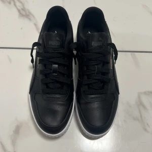 Svarta Puma sneakers med vit sula - Snygga svarta sneakers från Puma med klassisk design och vit sula. Skorna har snörning, rund tå och är tillverkade i skinnliknande material. Puma-logga i brons på sidan och bak på hälen. Perfekta för dig som gillar stilrena och sportiga skor.