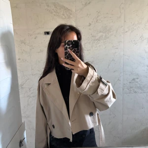 Beige croppad trenchcoat från Zara - Säljer en beige croppad trenchcoat från Zara med breda slag, svarta knappar och coola detaljer vid ärmsluten. Jackan har en loose passform och är tillverkad i bomullsmaterial. Perfekt för dig som gillar trendiga och avslappnade looks.