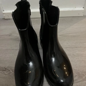 Svarta boots från Tommy Hilfiger - Snygga svarta boots i glansigt syntetmaterial från Tommy Hilfiger. De har bruna detaljer vid ankeln och ett guldfärgat spänne på sidan. Perfekta för regniga dagar och ger en stilren look till din outfit.