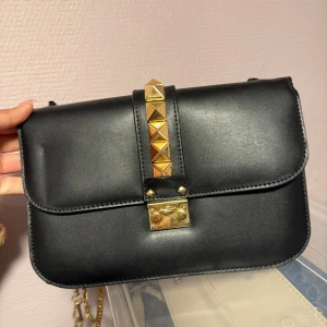 Svart axelväska med gulddetaljer - Snygg svart axelväska i skinn med guldfärgad kedja och coola pyramidformade nitar på framsidan. Väskan har ett stilrent, rektangulärt format och ett guldfärgat lås framtill. Perfekt för dig som gillar edgy detaljer och vill ha en väska som sticker ut.