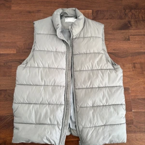 Ljusgrå dunväst från ASOS DESIGN - Snygg ljusgrå dunväst från ASOS DESIGN med quiltad design och hög krage. Västen har dragkedja framtill och är perfekt för lager-på-lager-stil. Enkel och clean look som passar till många outfits. Storleken är M men passar bäst på L. 