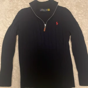Mörkblå kabelstickad tröja Polo Ralph Lauren - Kabelstickad mörkblå tröja från Polo Ralph Lauren med halv dragkedja och klassisk röd logga på bröstet. Tröjan har hög krage, lång ärm och ribbade muddar. Perfekt för dig som gillar stilren och tidlös design.